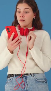 Kırmızı akıllı telefon ve kulaklıkla düşünceli bir kadın. Mavi arka planda kotlu ve süveterli sıradan tarz, müzikle ilgili bir şeyi yansıtıyor ya da karar veriyor gibi görünüyor. Dikey 