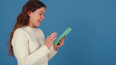 Beyaz bir kadın, elinde yeşil bir tablet tutarken ve gülümserken görülmüş. Günlük beyaz süveter, mavi arka plan ve temiz fotokopi alanı burayı modern ve minimal teknoloji sahnesi yapar. 