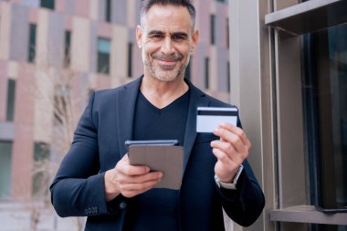 Elinde kredi kartı ve tablet tutarken kameraya gülümseyen beyaz bir adam. Şehir ortamında ayakta durmak, modern teknolojiyi temsil etmek, online alışveriş ve finansal güven. 