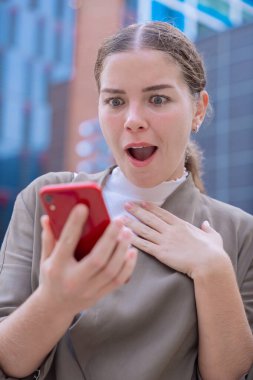 Kafkasyalı kadın şok olmuş ve şaşırmış bir ifadeyle akıllı telefonuna bakıyor. Beklenmedik haberler, sosyal medya tepkisi ve modern binalarla kentsel bir çevrede teknoloji kavramı. Dikey