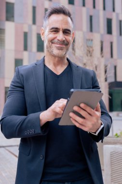 Modern bir ofis binasının dışında tablet üzerinde çalışan kendinden emin bir iş adamı. Kameraya bakıyorum. Kentsel ortam iş, teknoloji, üretkenlik ve profesyonel yaşam tarzı temaları için idealdir