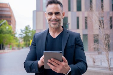 Modern bir ofis binasının dışında tablet üzerinde çalışan kendinden emin bir iş adamı. Kameraya bakıyorum. Kentsel ortam iş, teknoloji, üretkenlik ve profesyonel yaşam tarzı temaları için idealdir