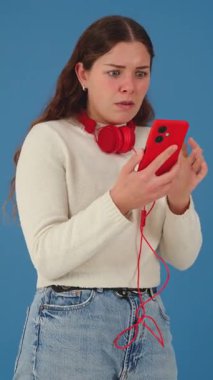 Akıllı telefonuyla video izleyen beyaz bir kadının dikey portresi. Kulaklık takıyor ve günlük giysiler giyiyor, temiz mavi arka planda dijital bir anın tadını çıkarıyor. Dikey