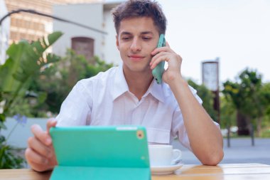 Kafkasyalı genç işadamı açık kafede birden fazla iş yapıyor, tablet kullanırken telefonla konuşuyor. Şehir yeşilliği ile çevrili, iş için ideal, uzak çalışma ve dijital yaşam tarzı kavramları. 