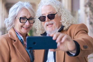 Kafkasyalı mutlu çift akıllı telefonuyla selfie çekiyor. Güneş gözlüğü ve ceket giyerek, parlak bir şekilde gülümserler. Açık havada aşk, neşe, bağlantı ve modern yaşam tarzı kavramı. 