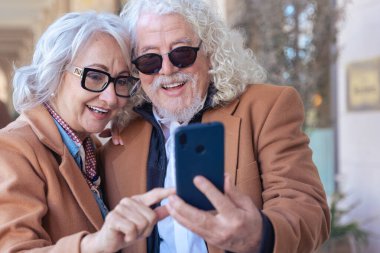 Gri saçlı ve gözlüklü yaşlı çift açık havada bir akıllı telefonu paylaşıyor. Gülüyorlar ve teknolojinin tadını çıkarıyorlar. Zarif bir şehir ortamında oturuyorlar. Kahverengi paltolarını giyip mutlu görünüyorlar..