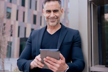 Modern bir ofis binasının dışında kameraya bakarak tablet üzerinde çalışan kendinden emin bir iş adamı. Kentsel ortam iş, teknoloji, üretkenlik ve profesyonel yaşam tarzı temaları için idealdir