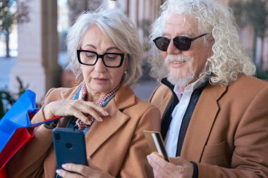Açık havada alışveriş yaparken ellerinde akıllı telefon ve kredi kartı tutan mutlu Kafkasyalı yaşlı çift. Şık ve neşeli, online alışveriş, emeklilik yaşam tarzı ve dijital ödeme gibi kavramlar için mükemmel.. 