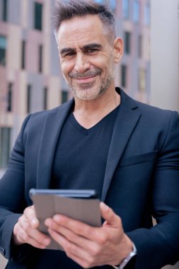 Modern bir ofis binasının önünde kameraya bakarak tablet üzerinde çalışan kendinden emin bir iş adamı. İş, teknoloji, üretkenlik ve mesleki yaşam tarzı temaları için ideal kentsel ortam. Dikey