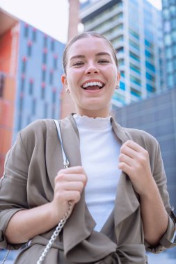 Genç beyaz iş kadını kameraya ışıltılı bir gülümsemeyle bakıyor. Kendine güven, mutluluk ve pozitiflik yayarak, onu yaşam tarzı, moda ve kişisel markalaşma konuları için ideal bir imaj haline getiriyor.