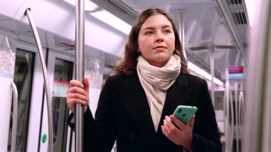Siyah paltolu ve bej eşarplı genç bir kadın modern bir metro treninin içinde elinde akıllı bir telefonla metal bir direği kavrıyor. İşe gidip gelirken odaklanmış görünüyor. Elinde şık beyaz bir çanta var.. 