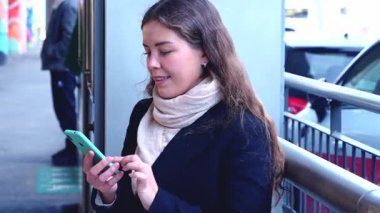 Paltolu ve fularlı genç bir kadın açık havada, toplu taşıma araçlarının yanında akıllı telefonunu kullanırken gülümsüyor. Telefonuyla meşgul görünüyor, rahat ve modern kentsel bir anın tadını çıkarıyor.. 