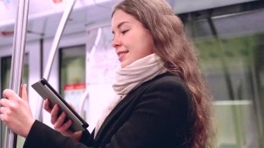 Modern bir metro treninde dururken siyah paltolu ve bej eşarplı şık bir kadın bir e-okuyucuda okuyor. Yolda giderken bir direğe tutunarak dengede duruyor.. 