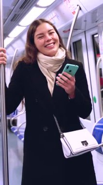 Trenle seyahat ederken ayakta duran akıllı telefonu kullanan beyaz bir kadın. Güzel, genç, gülümseyen bir kadın toplu taşıma aracı kullanıyor, kameraya bakıyor.