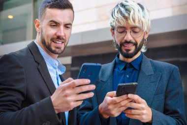 Takım elbiseli iki genç iş adamı şehirde dikilirken akıllı telefonlarını kullanıyor. Modern iletişim ve profesyonel dijital yaşam tarzını yansıtan odaklanmış ve meşgul görünüyorlar.. 