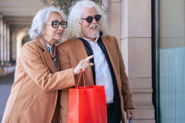 Gülümseyen yaşlı bir çift ellerinde alışveriş torbalarıyla dışarıda yürürken kadın bir mağazanın camını işaret ediyor. İkisi de kahverengi paltolu ve güneş gözlüklü. Keyifli ve rahat bir alışveriş gününün tadını çıkarıyorlar.. 
