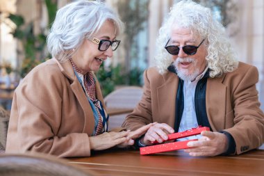 Dışarıda bir kafede partnerinden hediye alan son sınıf öğrencisi heyecanlı bir kadın. Yaşlı çift, bu görüntüyü aşk, sürpriz ve ilişki temaları için mükemmel kılan neşeli ve sevgi dolu bir anı paylaşıyor.