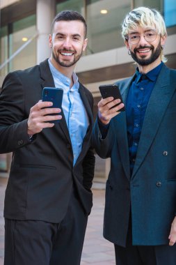 İki neşeli, kendine güvenen iş adamının portresi, gülümseyen ve akıllı telefonlar tutan, resmi kıyafetler giyen, modern ofis binalarının yanında duran, kameraya olumlu bakan.