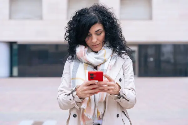 Şehir ortamında, paltosu ve atkısı olan, kırmızı akıllı telefonunu kullanan mutlu beyaz kadın. Gülümsüyor ve rahatlıyor. Soğuk bir günde sıradan bir anın tadını çıkarıyor.. 