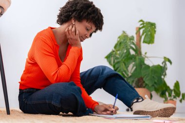 Turuncu kazaklı Afro-Amerikalı kadın yere oturmuş deftere bir şeyler yazıyor. Rahat, odaklanmış ve yaratıcı bir atmosferi olan doğal ışık ve yeşil bitkiyle huzurlu bir iç mekan sahnesi..