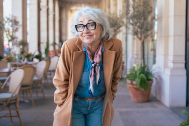 Mutlu Kafkasyalı kadın açık bir kafede güven içinde poz veriyor. Kahverengi bir ceket, eşarp ve gözlük giyerek, gün ışığında zarafet, deneyim ve zamansız moda ifade eder.. 