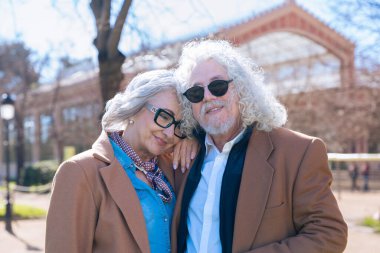 Gri saçlı ve gözlüklü yaşlı bir çift kameraya gülümsüyor. Adam güneş gözlüğü takıyor ve ikisi de zarif bir şekilde kahverengi paltolar giyip güneşli bir günün tadını çıkarıyor.. 