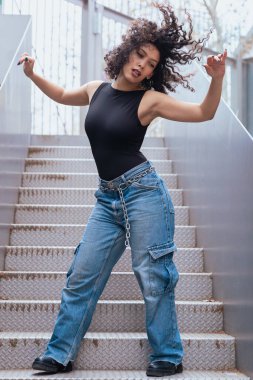 Kıvırcık saçlı, pembe ceketli, şehir ortamında hareket ve enerji ifade eden Afrikalı Amerikalı dansçı. Modern moda, çağdaş dans ve sanatsal performans.