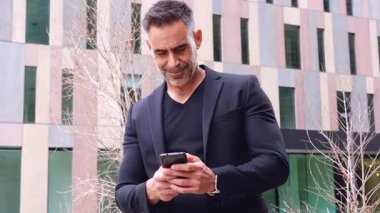 Modern bir binanın önünde duran siyah kıyafetli şık bir işadamı akıllı telefonuna bakıyor. Temiz, mimari çizgileri olan kentsel bir ortamda odaklanmış ve profesyonel görünüyor.. 