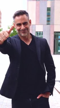 Siyahlar giymiş şık bir adam modern bir binanın önünde duruyor, kendine güvenerek gülümsüyor ve baş parmak işareti yapıyor. Şehir ortamında pozitif vücut dili ve poz. Dikey