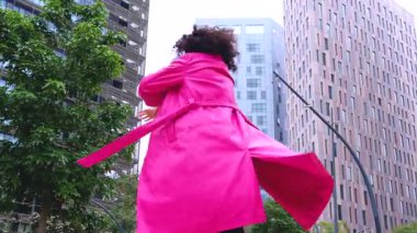 Pembe palto giyen kıvırcık saçlı Afrikalı Amerikalı dansçı şehir ortamında hareketlerini ve enerjilerini ifade ediyor. Modern moda, çağdaş dans ve sanatsal performans şehrin arka planına karşı.