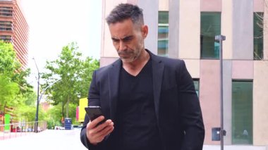 Siyah kıyafetli bir işadamı, bir şehir merkezinde, elinde akıllı bir telefonla kameraya bakıyor. Kendine güvenen ifade, modern ortam ve sıradan ama profesyonel görünüş.. 