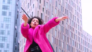 Pembe palto giyen kıvırcık saçlı Afrikalı Amerikalı dansçı şehir ortamında hareketlerini ve enerjilerini ifade ediyor. Modern moda, çağdaş dans ve sanatsal performans şehrin arka planına karşı.