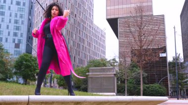 Pembe palto giyen kıvırcık saçlı Afrikalı Amerikalı dansçı şehir ortamında hareketlerini ve enerjilerini ifade ediyor. Modern moda, çağdaş dans ve sanatsal performans şehrin arka planına karşı.