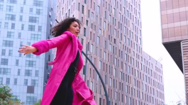 Pembe palto giyen kıvırcık saçlı Afrikalı Amerikalı dansçı şehir ortamında hareketlerini ve enerjilerini ifade ediyor. Modern moda, çağdaş dans ve sanatsal performans şehrin arka planına karşı.