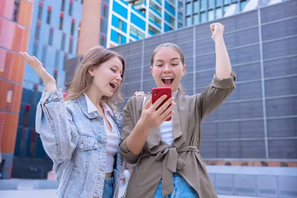 İnternetteki bir iddiayı kazandıktan sonra dışarıda kutlama yapan iki beyaz kadın. Akıllı bir telefona bakarken heyecanla kollarını kaldırıyorlar. Neşe, başarı ve dijital yaşam biçimi eylem sırasında yakalanır. 