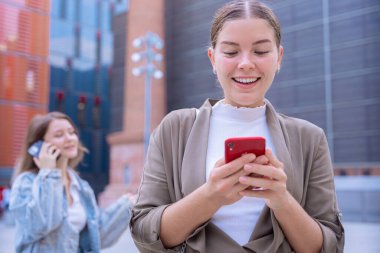 Modern bir şehirde akıllı telefon kullanan iki beyaz kadın var. Bir mesajlaşma, bir telefonda konuşma, sıradan kıyafetler, arka planda yüksek binalar. Bağlı, dijital ve kentsel yaşam tarzı. 