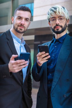 İki neşeli, kendine güvenen iş adamının portresi, gülümseyen ve akıllı telefonlar tutan, resmi kıyafetler giyen, modern ofis binalarının yanında duran, kameraya olumlu bakan.