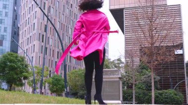 Pembe palto giyen kıvırcık saçlı Afrikalı Amerikalı dansçı şehir ortamında hareketlerini ve enerjilerini ifade ediyor. Modern moda, çağdaş dans ve sanatsal performans şehrin arka planına karşı.