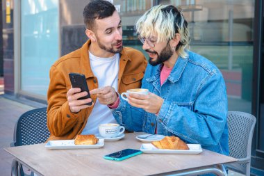 Gülümseyen adam elinde kahve ve pastayla açık kafede otururken diğer bir kişi de POS terminali ile kart ödemesini işliyor. Sıradan kentsel ortam, doğal ışık, rahat ve modern yaşam tarzı anı. 