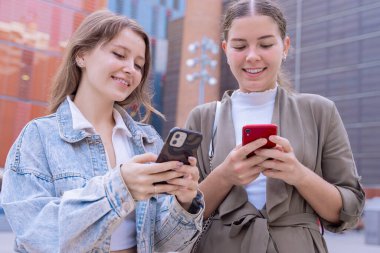 İki beyaz kadın modern bir şehirde akıllı telefon kullanıyor. Biri mesajlaşıyor, diğeri telefonla konuşuyor. Sıradan kıyafetler, arka planda yüksek binalar. Bağlı, dijital .