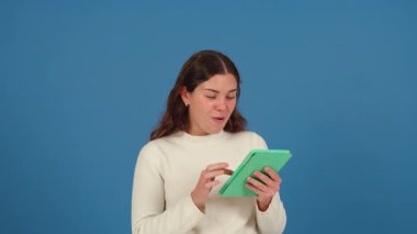 Beyaz bir kadın, elinde yeşil bir tablet tutarken ve gülümserken görülmüş. Günlük beyaz süveter, mavi arka plan ve temiz fotokopi alanı burayı modern ve minimal teknoloji sahnesi yapar. 