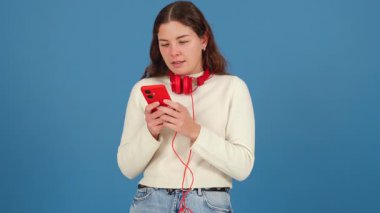 Kırmızı kulaklıklı ve akıllı telefonlu beyaz bir kadın düşünceli görünüyor. Sıradan bir süveter ve kot pantolon giyerek, mavi bir arka plana karşı duruyor, modern teknoloji bağlamında şüphe veya yansımasını ifade ediyor. 