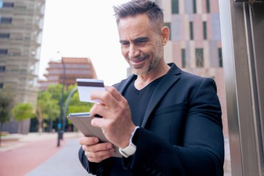 Kafkasyalı yetişkin bir adam tablet kullanıyor ve elinde online ödeme yapmak için bir kredi kartı tutuyor. Modern finans, alışveriş ve dijital yaşam tarzını temsil eden kentsel bir ortamda odaklanmış ifade 