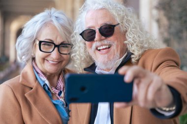 Kafkasyalı mutlu çift akıllı telefonuyla selfie çekiyor. Güneş gözlüğü ve ceket giyerek, parlak bir şekilde gülümserler. Açık havada aşk, neşe, bağlantı ve modern yaşam tarzı kavramı. 