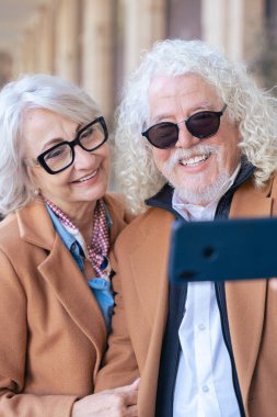 Kafkasyalı mutlu çift akıllı telefonuyla selfie çekiyor. Güneş gözlüğü ve ceket giyerek, parlak bir şekilde gülümserler. Açık havada aşk, neşe, bağlantı ve modern yaşam tarzı kavramı. 