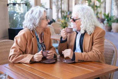 Gözlüklü ve kahverengi paltolu yaşlı bir çift açık hava kafesinde kahvenin tadını çıkarıyor. Sıcak bir anı paylaşıyorlar, gülümsüyorlar ve gündüzleri huzurlu, rahat bir atmosferde rahatlıyorlar..
