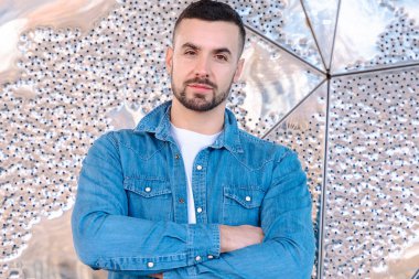 Kolları çaprazlanmış kot gömlek giyen, parlak ve soyut bir arka planın önünde duran ciddi bir gencin portresi. Kentsel tavır ve modern erkeksi güven. 