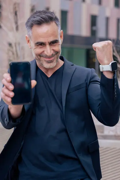 Siyahlı mutlu işadamı online bahsi kazanmasını kutluyor. Akıllı telefonunu kaldırıp zaferle yumruğunu kaldırıyor. O, neşe ve heyecan dolu modern bir şehir bölgesinde duruyor. Dikey