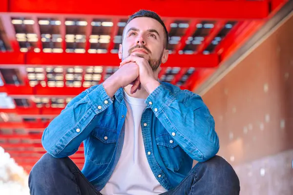 Kot gömlekli düşünceli bir adam çarpıcı bir kırmızı kentsel yapının altında oturuyor, güçlü geometrik kontrastlı modern şehir ortamında yansıma, yaşam tarzı ve kişisel düşünce aktarıyor. 