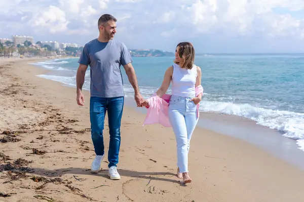 Kumsalda çıplak ayakla yürüyen, el ele tutuşan ve tatilde gülümseyen beyaz bir çift. Okyanus dalgaları, kum ve arka planda parlak bir gökyüzü olan sakin deniz kenarı anı. Boşluğu kopyala. 